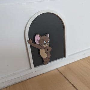 May include: A decorative wall decal featuring a brown cartoon mouse character leaning against the edge of a black arched doorway. The arch has a white frame and the mouse has large pink-lined ears, a tan belly, and a confident facial expression.