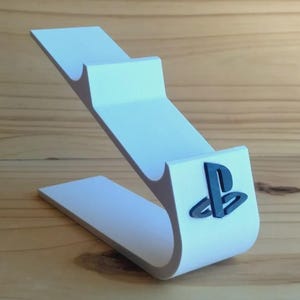 May include: A white, curved controller stand featuring a tiered design with two distinct cradles for holding a gaming controller. The front of the stand displays a dark blue, raised PlayStation logo emblem attached to the smooth, matte surface.