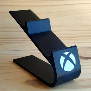 May include: A black controller stand featuring a curved base and an angled support arm with a notched cradle. The front of the stand displays a circular white Xbox logo emblem. The structure is designed to hold a gaming controller securely in place.