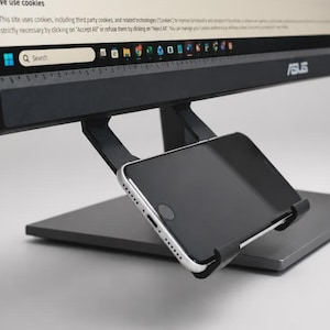 May include: A black smartphone mount attached to the side of an ASUS computer monitor. The mount features a black plastic arm and a cradle holding a silver and black smartphone in a horizontal position, with the screen facing outward.