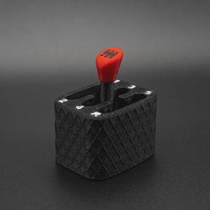 May include: A black gear shifter base with a diamond-textured exterior and white gear position markings including 1, 2, 3, 4, 5, and R. A red gear shift knob with a black gear pattern sits in the centre of the rectangular black housing.