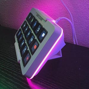 May include: A white macro keypad with the text AMPLIGAME printed on the top edge. The device features a grid of black keys with backlit icons and a vibrant pink LED light strip running along the side. A white cable is connected to the top port.