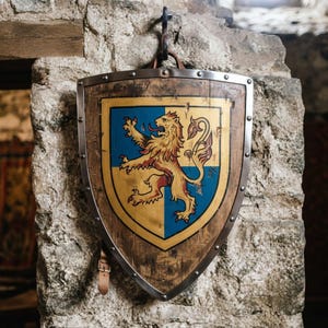 May include: A wooden shield featuring a metal rim and a central painted emblem of a golden lion rampant on a blue and yellow field. The shield has a pointed base, a leather strap at the bottom, and a metal hook at the top for wall mounting.