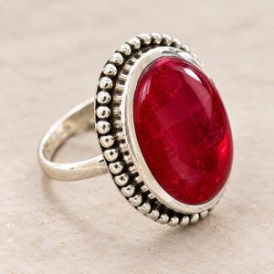 May include: A silver ring featuring a large, oval-shaped, deep red gemstone set in a polished metal bezel. The setting is surrounded by a decorative border of small, rounded silver beads, creating a textured frame around the central stone.
