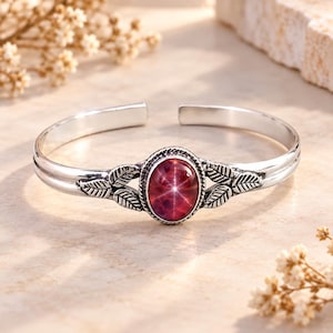 Puede incluir: Una pulsera de tipo brazalete en tono plateado con una piedra cabujón ovalada de color rojo intenso que presenta un reflejo de luz en forma de estrella en el centro. La banda metálica tiene pequeños motivos de hojas en relieve a los lados.