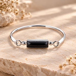 Op de afbeelding: Een zilverkleurige armband met een centrale rechthoekige zwarte edelsteen. De armband heeft een gladde, gepolijste metalen band die aan beide zijden met de zetting is verbonden via kleine ronde ringetjes en bolvormige metalen accenten.