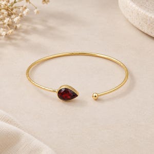 Puede incluir: Una pulsera abierta de tono dorado que presenta una piedra preciosa de color rojo oscuro facetada en forma de pera en un extremo y una pequeña esfera dorada pulida en el otro. La fina banda de metal tiene una textura martillada sutil.