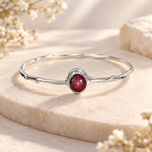 Puede incluir: Una pulsera rígida de plata con una banda texturizada similar a una rama y una piedra preciosa central redonda de color rojo intenso con efecto estrella. El metal tiene un acabado pulido y el engaste de la piedra tiene un borde irregular.