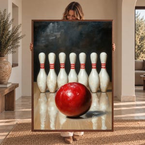 May include: A framed rectangular wall art piece featuring a painting of a glossy red bowling ball positioned in the foreground on a polished wooden lane, with seven white bowling pins with red stripes standing in a row against a dark background.