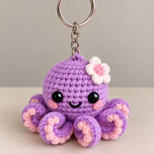 May include: A purple crochet octopus keyring featuring a small white flower with a pink centre attached to its head. The octopus has black plastic eyes, pink stitched cheeks, a black stitched smile, and light pink undersides on its tentacles.