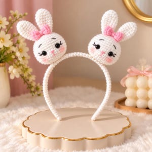 Pode incluir: Uma tiara de crochê branca com duas cabeças de coelho acolchoadas. Cada coelho tem orelhas longas, um pequeno laço rosa, olhos pretos, cílios bordados e bochechas rosadas. A tiara em si é coberta com pontos de crochê brancos.