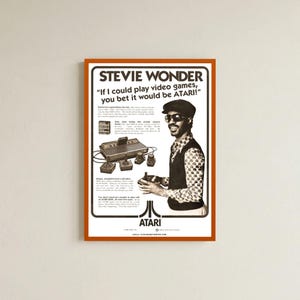 Puede incluir: Un póster publicitario vintage de Atari con Stevie Wonder usando una gorra y gafas de sol. El texto dice, Stevie Wonder, If I could play video games, you bet it would be ATARI! Incluye una consola Atari 2600 y joysticks.