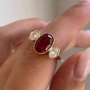 Può includere: Un anello in oro con una grande pietra centrale di rubino rosso intenso a taglio ovale, affiancata da due diamanti bianchi trasparenti più piccoli a taglio ovale. Le pietre sono incastonate in una montatura liscia su una fascia in oro lucido.