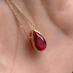 Pode incluir: Um colar de corrente dourada com um pingente de pedra preciosa vermelha lapidada em forma de pera, engastado em uma moldura dourada. O pingente pende de uma delicada corrente de elos dourados, exibindo uma cor carmesim e acabamento metálico.