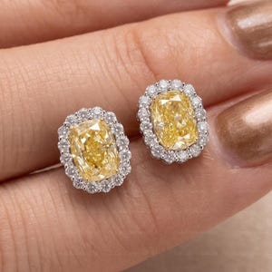 May include: A pair of stud earrings featuring a central cushion-cut yellow gemstone surrounded by a halo of small, round, clear white diamonds set in a polished silver-toned metal frame, creating a bright and reflective appearance.