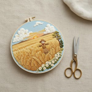 Puede incluir: Un bastidor de bordado circular que contiene un paisaje cosido de un campo de trigo dorado bajo un cielo azul claro con nubes blancas y un sol. Un pequeño espantapájaros se encuentra en el centro, rodeado de flores blancas y un árbol, con tijeras.