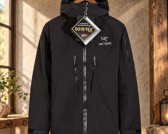 May include: A black Arc'teryx hooded jacket featuring a full-length front zip, two vertical chest pockets with zips, and a Gore-Tex product tag attached to the collar. The jacket has long sleeves with adjustable cuffs and a white logo on the chest.