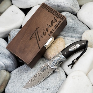 May include: A dark wood rectangular box with a silver metal latch, engraved with the text GROOMSMAN, Thomas, and 7-21-2026. Beside it lies a folding pocket knife with a patterned Damascus steel blade and a dark handle featuring a silver inlay.