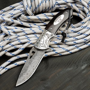 May include: A folding pocket knife with a Damascus steel blade featuring a wavy pattern and a dark wood handle. The handle includes a silver inlay engraved with the text DOMINIC BEST MAN 7.21.2026 and a black leather lanyard with a silver bead.