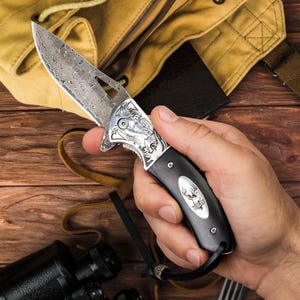 May include: A folding pocket knife with a Damascus steel blade featuring a wavy pattern and a cutout. The handle is black with a silver oval inlay displaying a deer head graphic and the text THOMAS. The bolster features an engraved animal motif.
