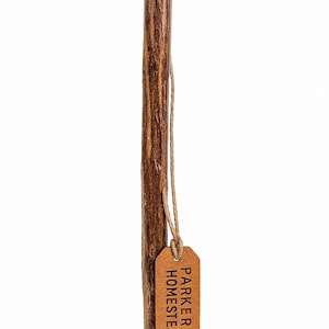 May include: A brown wooden walking stick with a textured surface. A small, brown leather tag is attached with twine, displaying the text "PARKER HOMESTEAD". The walking stick is a natural color and appears sturdy.
