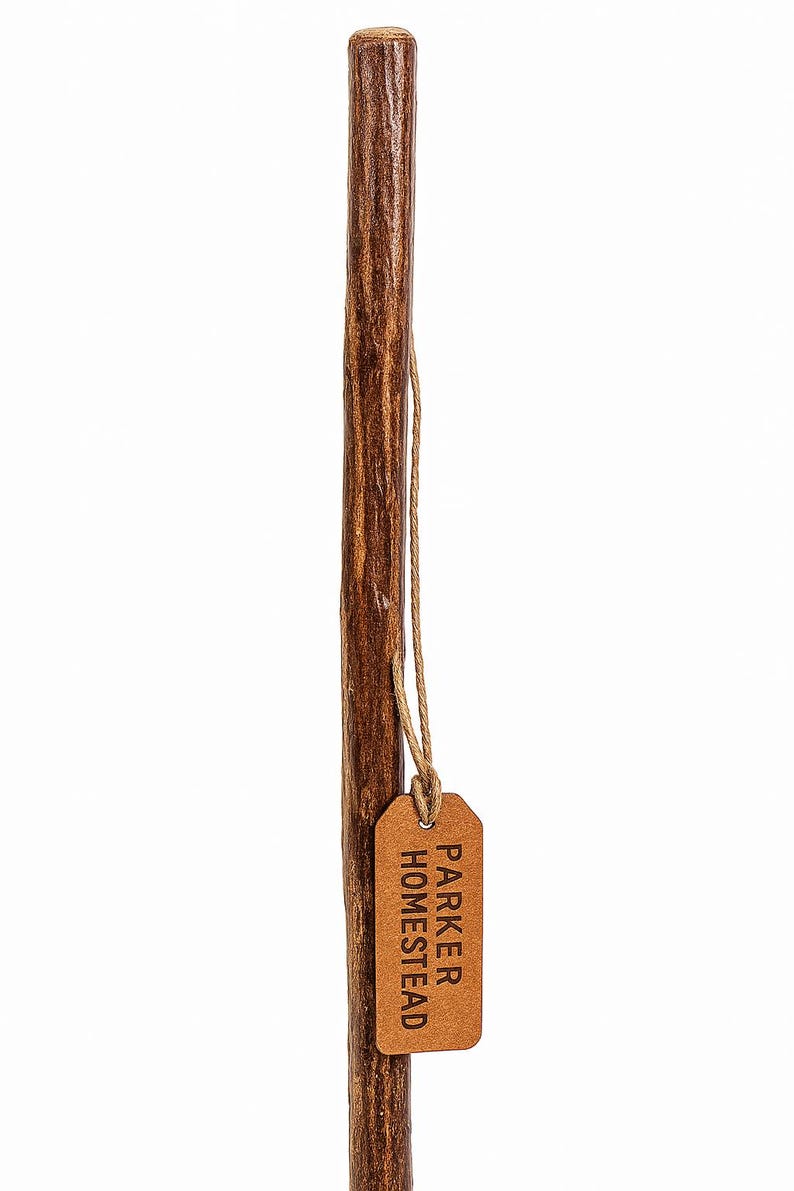 May include: A brown wooden walking stick with a textured surface. A small, brown leather tag is attached with twine, displaying the text "PARKER HOMESTEAD". The walking stick is a natural color and appears sturdy.