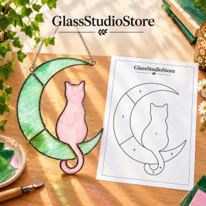 May include: A stained glass suncatcher featuring a light pink cat sitting on a crescent moon made of light green glass segments. The piece hangs from a metal chain and is accompanied by a printed paper pattern labeled GlassStudioStore.