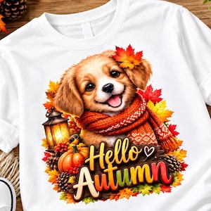 Könnte beinhalten: Ein weißes T-Shirt mit einer Grafik eines Welpen, der einen orangefarbenen Strickschal trägt und ein Ahornblatt auf dem Kopf hat. Das Motiv zeigt eine leuchtende Laterne, einen Kürbis, Tannenzapfen und herbstliche Blätter mit dem Text Hello Autumn.