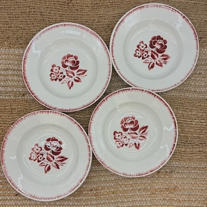 Peut inclure: Un ensemble de quatre assiettes en céramique blanche, chacune ornée d'un motif floral rouge foncé au centre et d'une bordure à tirets rouge foncé sur le rebord. Les assiettes sont disposées en carré sur une surface brune texturée.