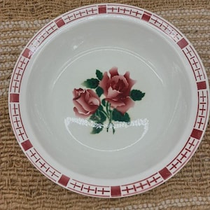 Peut inclure: Un bol rond en céramique blanche orné d'une illustration centrale de deux roses roses avec des feuilles vertes. Le bord est décoré d'un motif répétitif de carrés rouge foncé et de fines lignes formant une grille en croix.