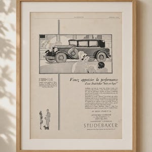 Könnte beinhalten: Eine gerahmte Vintage-Werbung für ein Studebaker-Automobil mit einer schwarz-weißen Illustration eines klassischen Wagens und Personen in Kleidung der 1920er Jahre. Der Text lautet "Venez apprécier la performance d'une Studebaker huit-en-ligne".