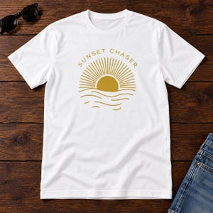 May include: A white short-sleeved t-shirt featuring a gold graphic of a sun setting over wavy lines with the text SUNSET CHASER arched above the sun. The design is centred on the chest of the shirt, which has a crew neckline and short sleeves.