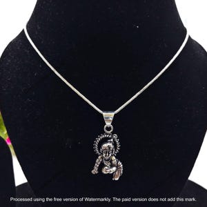 Könnte beinhalten: Eine silberfarbene Metallhalskette mit einer dünnen Kette und einem Anhänger, der eine kleine Figur in kriechender Position darstellt, umgeben von einem kreisförmigen, texturierten, heiligenscheinartigen Rahmen. Der Anhänger hängt an einer dreieckigen Öse.