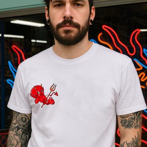 Puede incluir: Camiseta blanca con un diablo rojo de dibujos animados sosteniendo un tridente. El diablo está acostado. La camiseta es de manga corta y tiene cuello redondo. La persona que lleva la camiseta tiene tatuajes en los brazos.