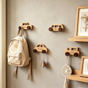 Puede incluir: Cuatro ganchos de pared de madera con forma de coche, cada uno con una rueda marrón oscuro y un nombre grabado en el cuerpo: Leo, Mia, Noah y Lily. Los ganchos están montados en una pared, con una mochila beige y una campana colgadas.
