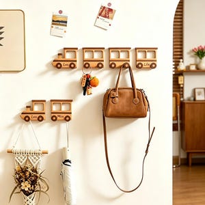 May include: A set of six wooden wall-mounted hooks shaped like train cars, featuring natural light wood tones and dark brown accents. Each hook displays a name including Leo, Mia, and Lila. The hooks are used to hang a brown handbag, keys, and an umbrella.