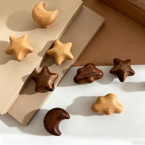 May include: A collection of wooden cabinet knobs in various shapes including stars, moons, and clouds. The knobs are finished in light and dark wood tones, featuring smooth, rounded edges and visible natural wood grain patterns throughout.