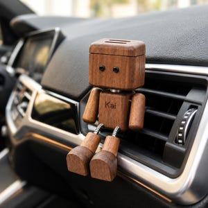 May include: A wooden robot-shaped car air freshener clip with a dark brown finish, featuring two small black screw eyes for eyes and the name Kai engraved on the chest. The figure has articulated arms and legs connected by silver springs.