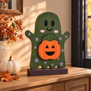 May include: A green wooden ghost cutout standing on a dark rectangular base. The ghost features black eyes and a mouth, pink cheeks, and purple stars. It holds an orange jack-o'-lantern bucket with a black handle and a carved face.