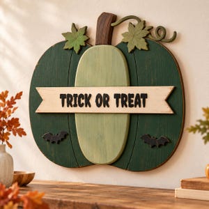 Pode incluir: Uma placa de parede de madeira em forma de abóbora pintada em tons de verde escuro e claro com um caule castanho e folhas verdes. Uma faixa horizontal creme no centro apresenta o texto preto TRICK OR TREAT, ladeado por dois morcegos pretos.