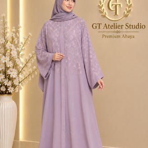 May include: A floor-length lavender abaya featuring a matching hijab, long flared sleeves, and a delicate gold floral pattern across the upper bodice. The garment includes a central vertical seam and is branded with the text GT Atelier Studio Premium Abaya.