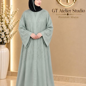 May include: A sage green abaya featuring long, wide sleeves and delicate tonal embroidery across the chest and sleeves. The garment is worn with a black hijab that has a white stripe detail. The background displays the text GT Atelier Studio.