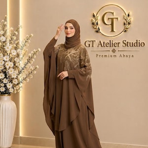 Puede incluir: Una abaya marrón sólida que presenta una silueta fluida y en capas con mangas largas y un hiyab a juego. El área superior del pecho está adornada con intrincados bordados en tono dorado. El texto GT Atelier Studio Premium Abaya es visible.