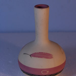May include: A small, white ceramic vase with a pink band and black geometric designs. The vase has a pink feather design painted on the front.