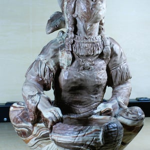 May include: A marbled brown and cream ceramic sculpture of a seated figure with braided hair, wearing a fringed tunic and a necklace with a central pendant. The figure is positioned with hands resting on a textured, woven-style ceramic basket.