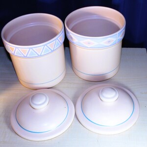 May include: Two ceramic canisters with lids, featuring a light pink base with blue and pink geometric designs. The lids are also light pink with a blue trim.
