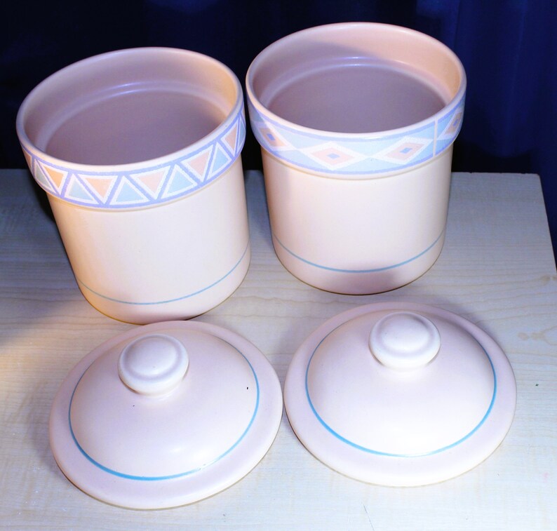 May include: Two ceramic canisters with lids, featuring a light pink base with blue and pink geometric designs. The lids are also light pink with a blue trim.
