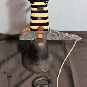 May include: A black and white striped lighthouse with a red roof and a black bell with a rope attached. The lighthouse is sitting on a gray base and the bell is on a black surface.