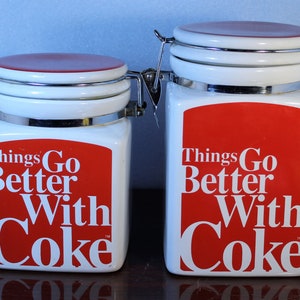 May include: Two white ceramic canisters with red lids and red and white lettering that reads "Things Go Better With Coke".