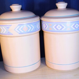 May include: Two ceramic canisters with lids, featuring a light pink base with a blue and white geometric pattern. The canisters are ideal for storing dry goods in the kitchen.